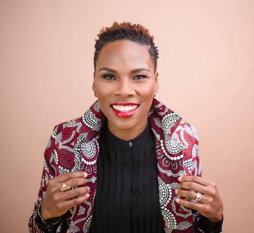 Luvvie in colorful pattern jacket smiling into camera in front of light pink solid background