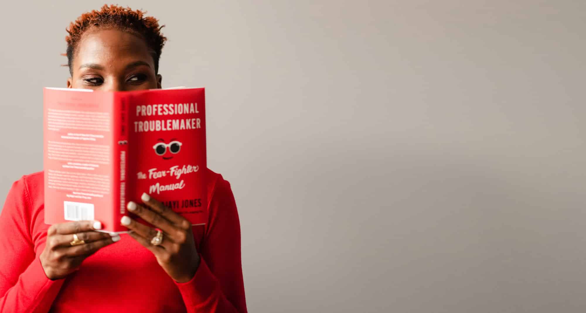 Luvvie in red shirt reading Professional Troublemaker book with red cover