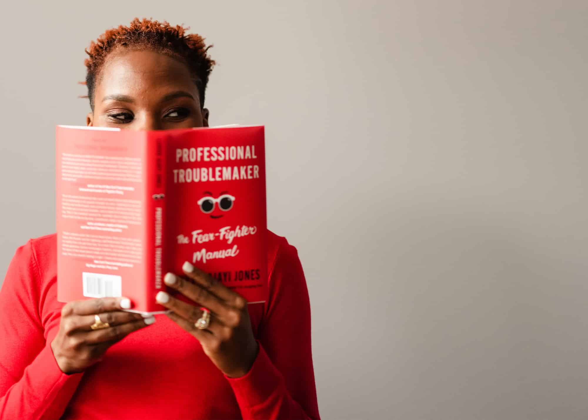 Luvvie in red shirt reading Professional Troublemaker book with red cover