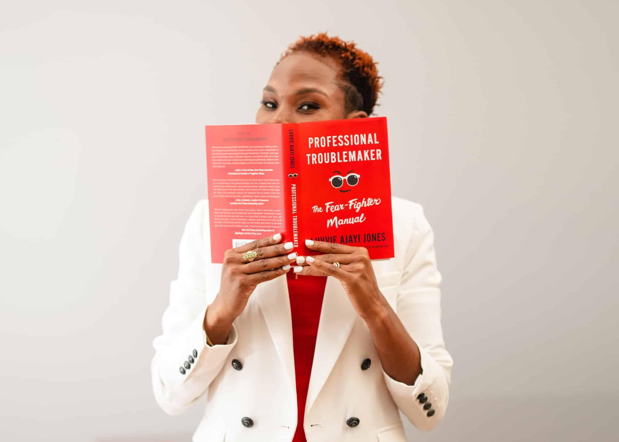 Luvvie in white blazer holding up and peaking over her book Professional Troublemaker