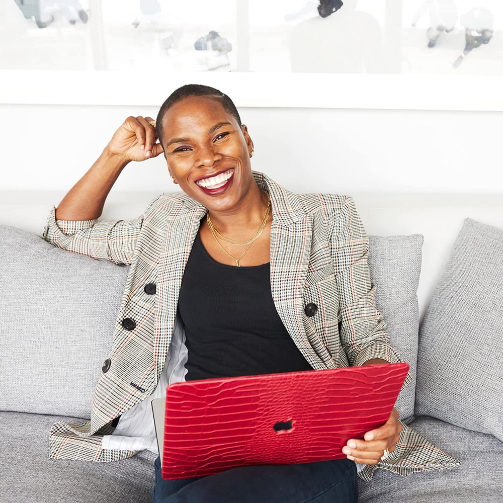 Luvvie leaning on her hand and smiling sitting on a couch with a red laptop on her lap