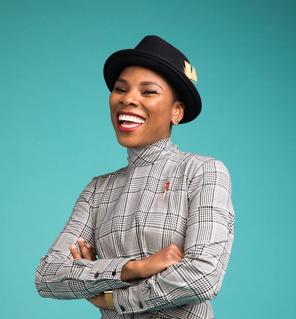 Luvvie smiling with arms crossed standing in front of colorful teal background wearing hat and long-sleeve shirt