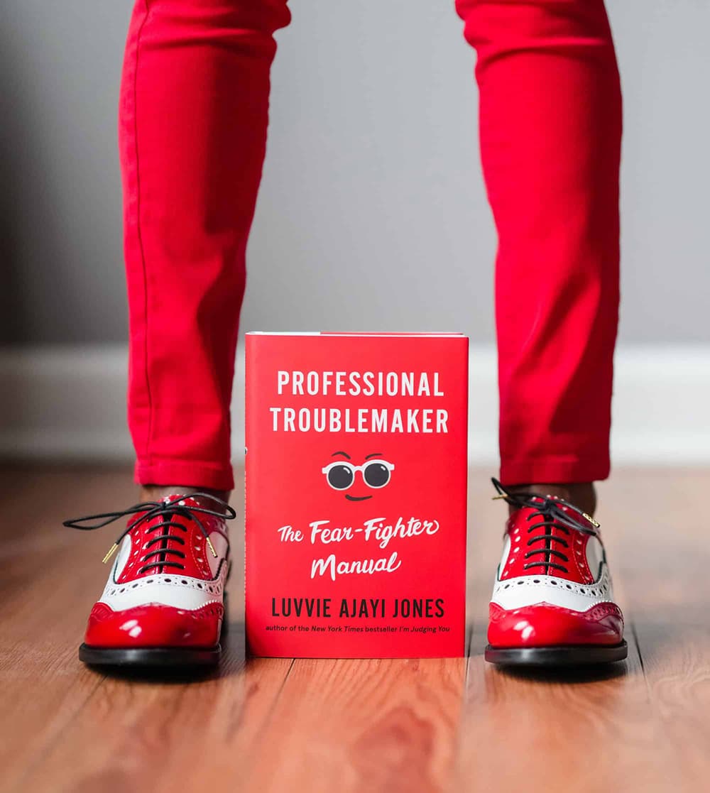 Close up of Professional Troublemaker book with red cover and person's legs in red pants and red shoes standing over it