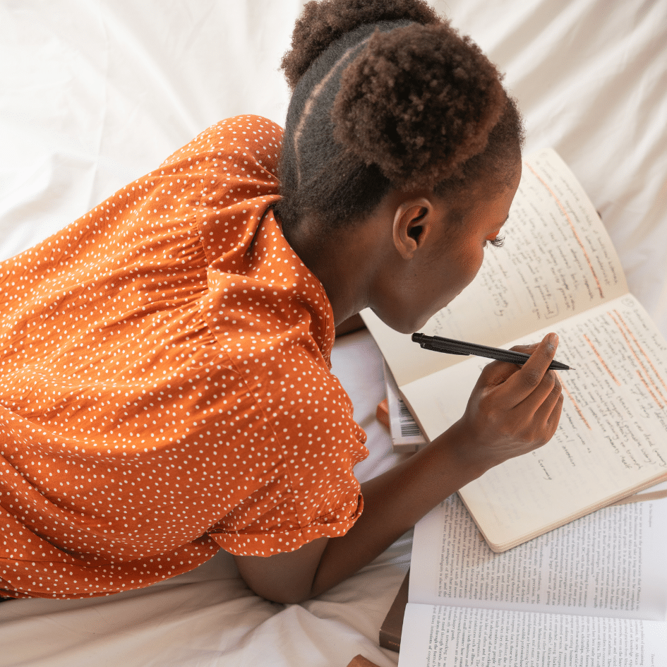 Aerial shot of girl in orange shirt lying on a bed writing in a notebook