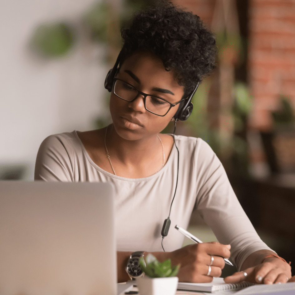 Woman wearing headphones looking at computer screen and writing in a notebook