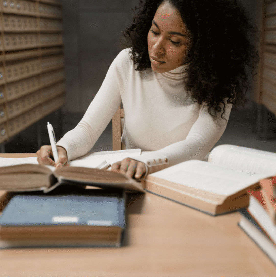 Woman in white long-sleeve turtleneck sweater writing surrounded by open books