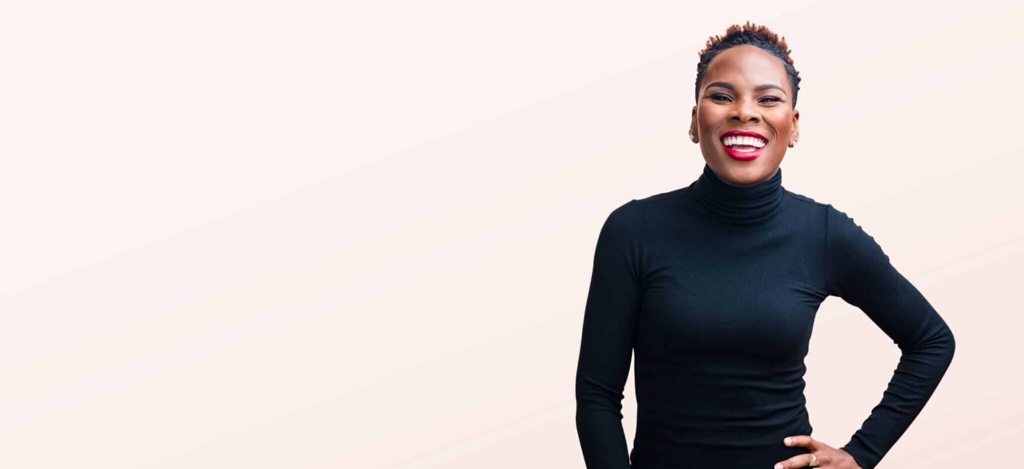 Luvvie in long-sleeve black turtleneck smiling with hand on her hip in front of solid light pink background