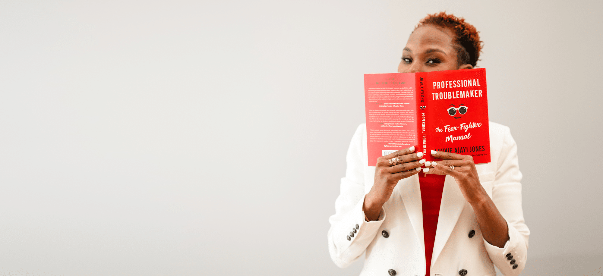 Luvvie in white blazer holding up and peaking over her book Professional Troublemaker