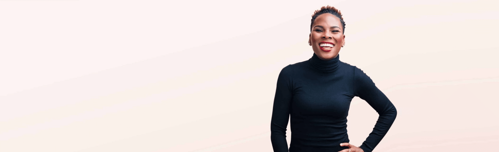 Luvvie in long-sleeve black turtleneck smiling with hand on her hip in front of solid light pink background